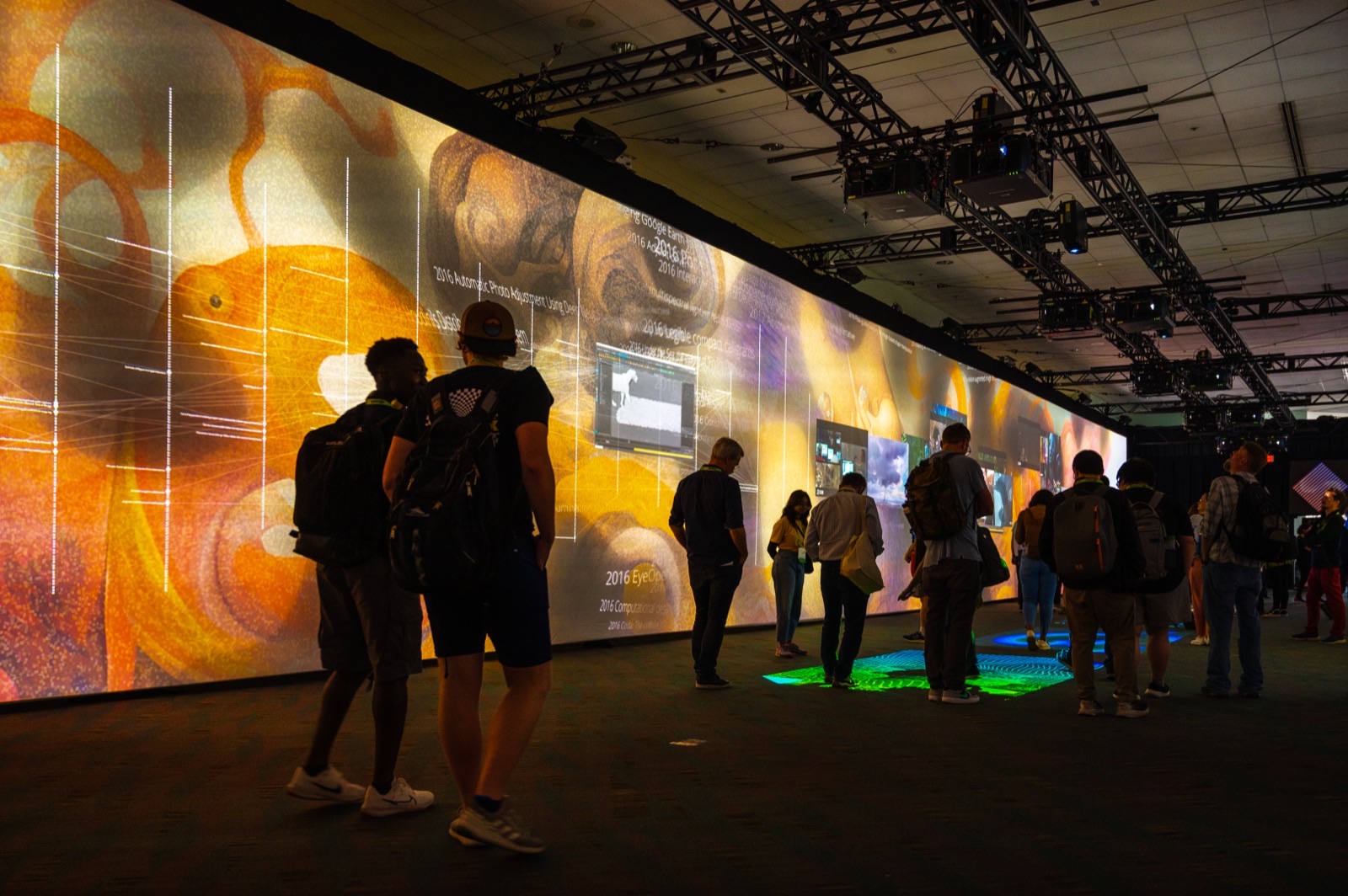 Visitors silhouetted against a wall of amber generative imagery threaded with paper titles from 2016