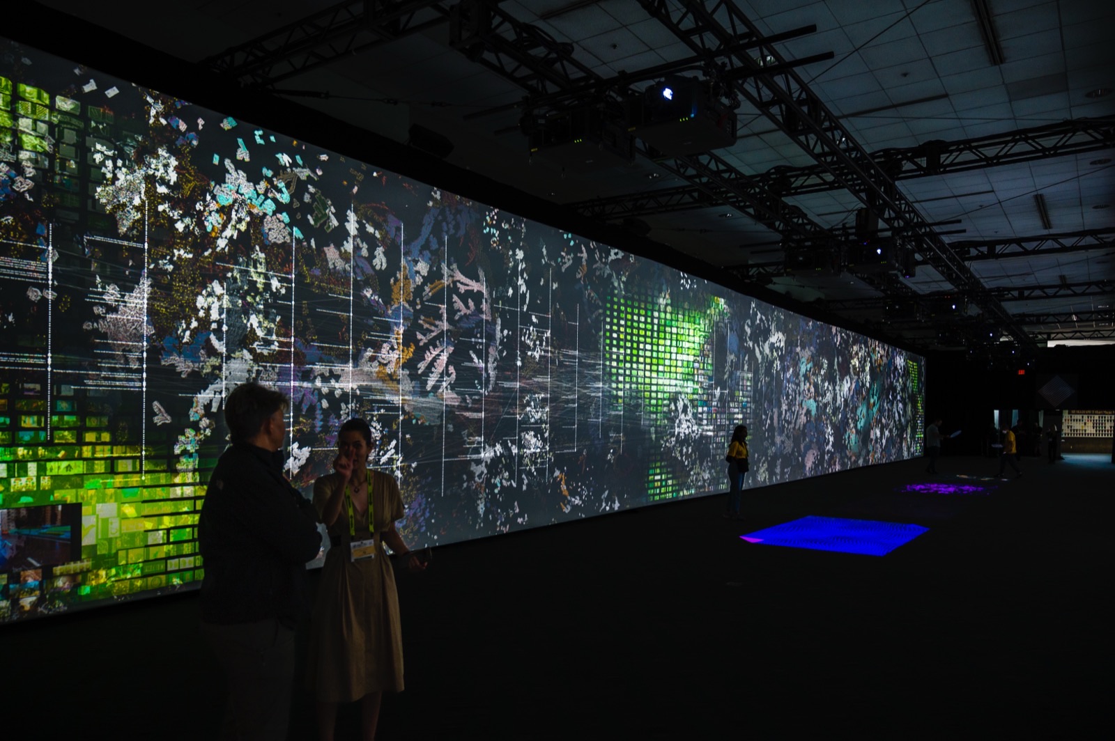 A long LED wall running the length of the room, dense with drifting thumbnails and text, with coloured interactive patches projected onto the floor beside it